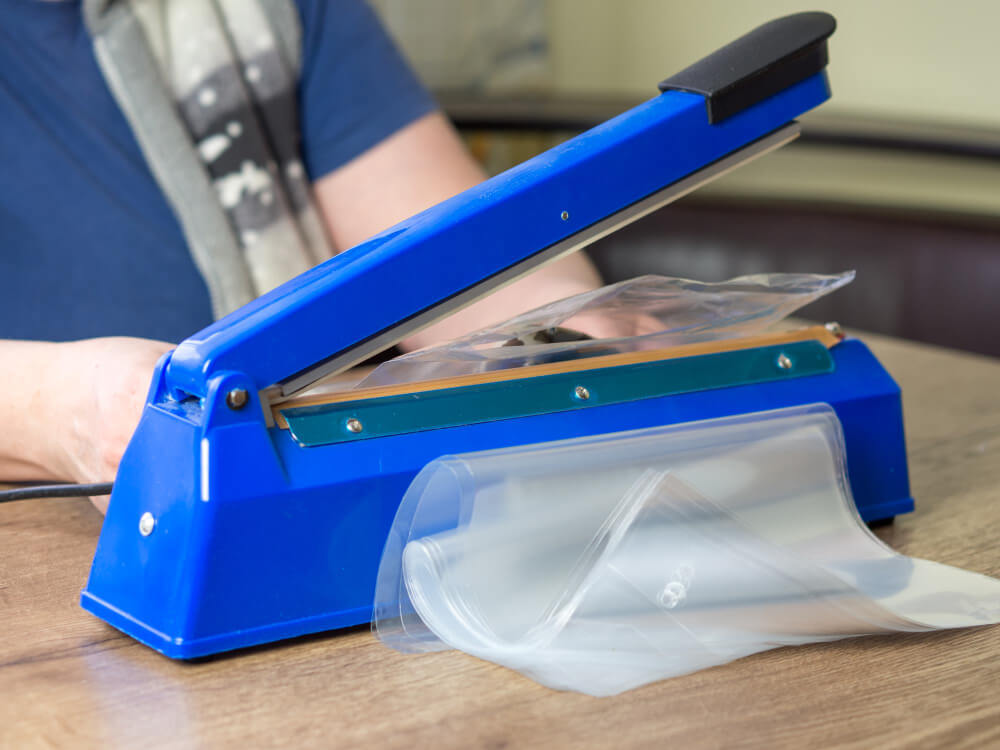 מכונת הלחמת שקיות מאמר | בתמונה: מכונת הלחמת שקיות ידנית בצבע כחול עם ערימת שקיות אטומות מהצד השני שלה